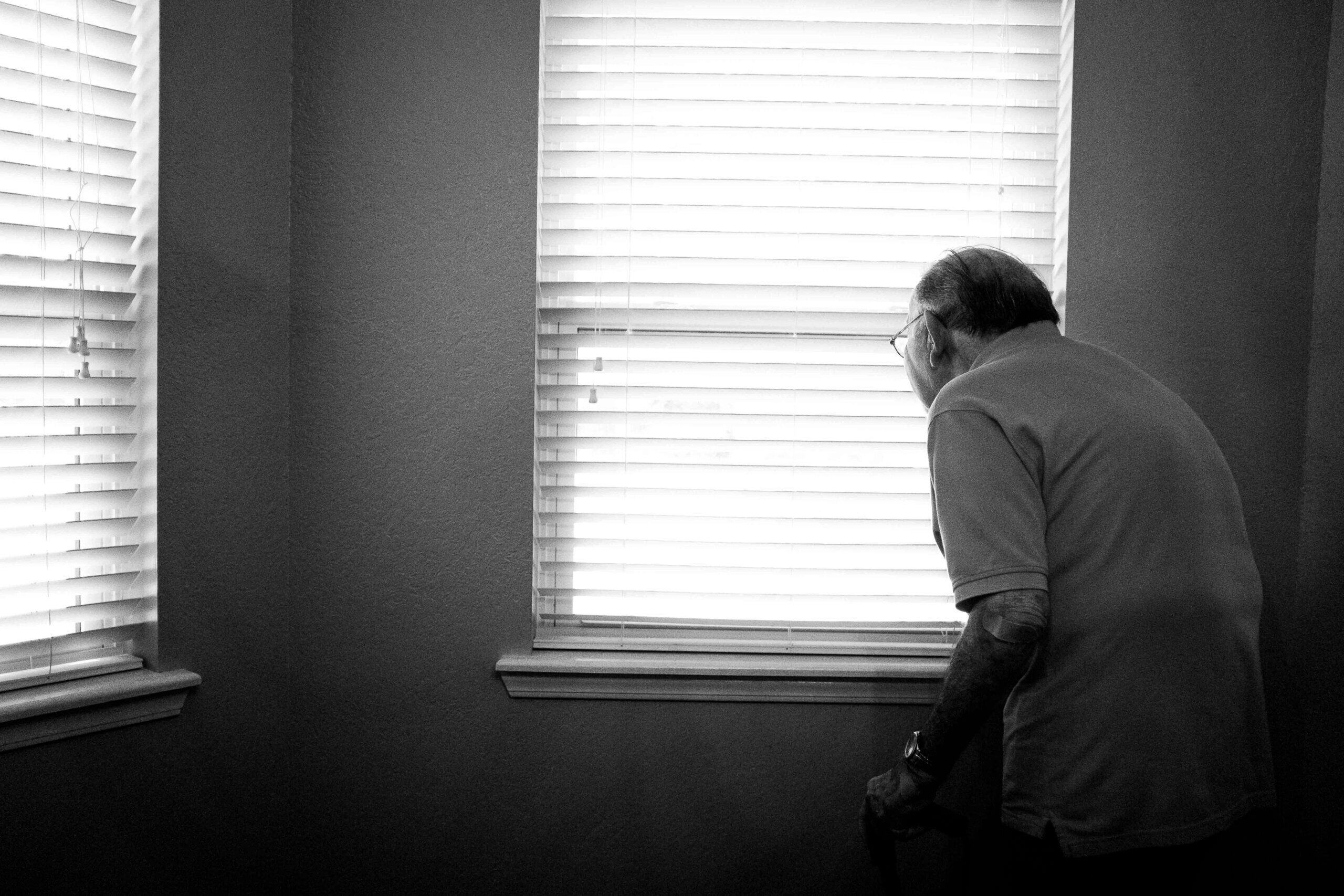 Pogrbljeni umirovljenik stoji u bijeloj sobi pored prozora i promatra okolinu.