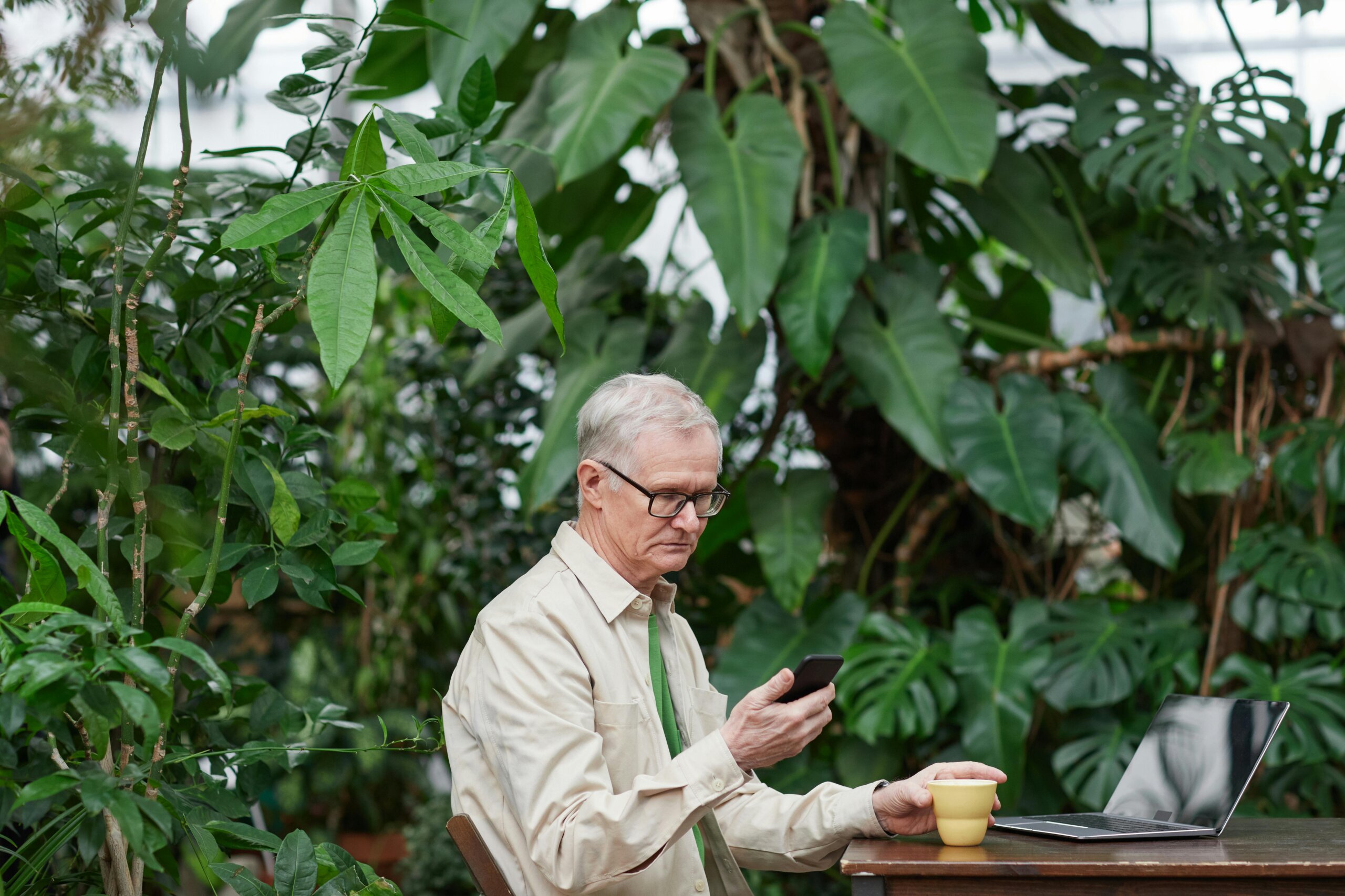 Umirovljenik u bež jakni sjedi za stolom i gleda u mobitel dok pije kavu.