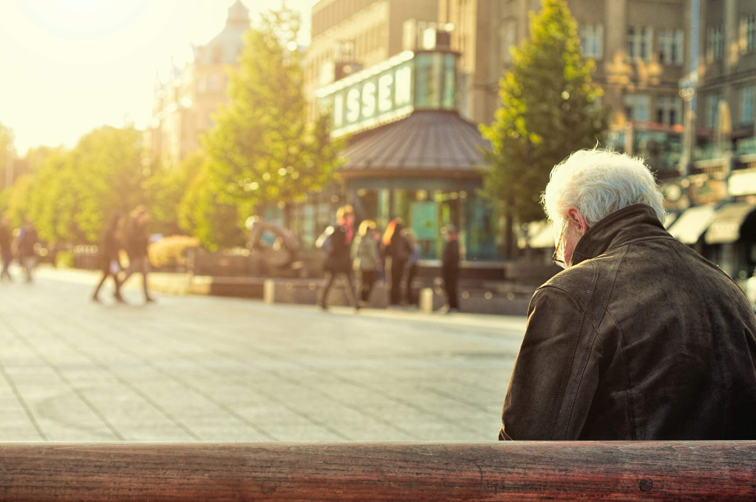 Umirovljenik u kožnoj jakni sjedi na smeđoj drvenoj klupi.