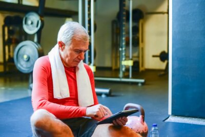 Umirovljenik sjedi na podu teretane nakon treninga i prati svoj program tjelovježbe na tabletu.