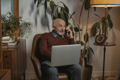 Umirovljenik telefonira dok na nogama drži prijenosno računalo (laptop).